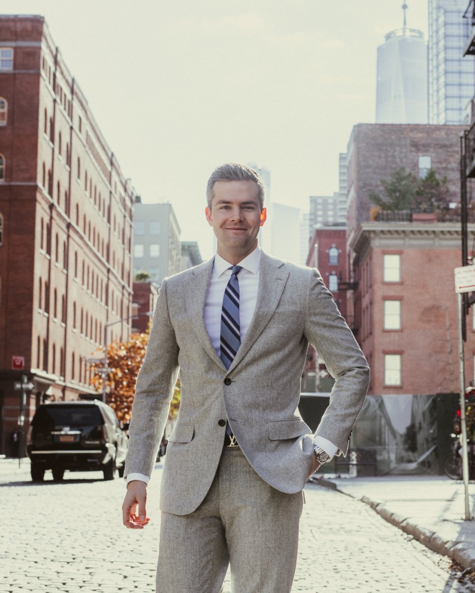 Ryan Serhant with a smile wearing a grey suit against a backdrop of a sunkissed New York City