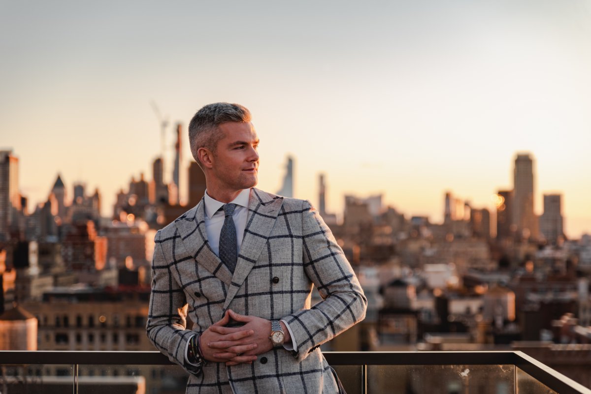 Ryan Serhant looking pensive wearing a windowpane grey suit against a cityscape skyline at dusk