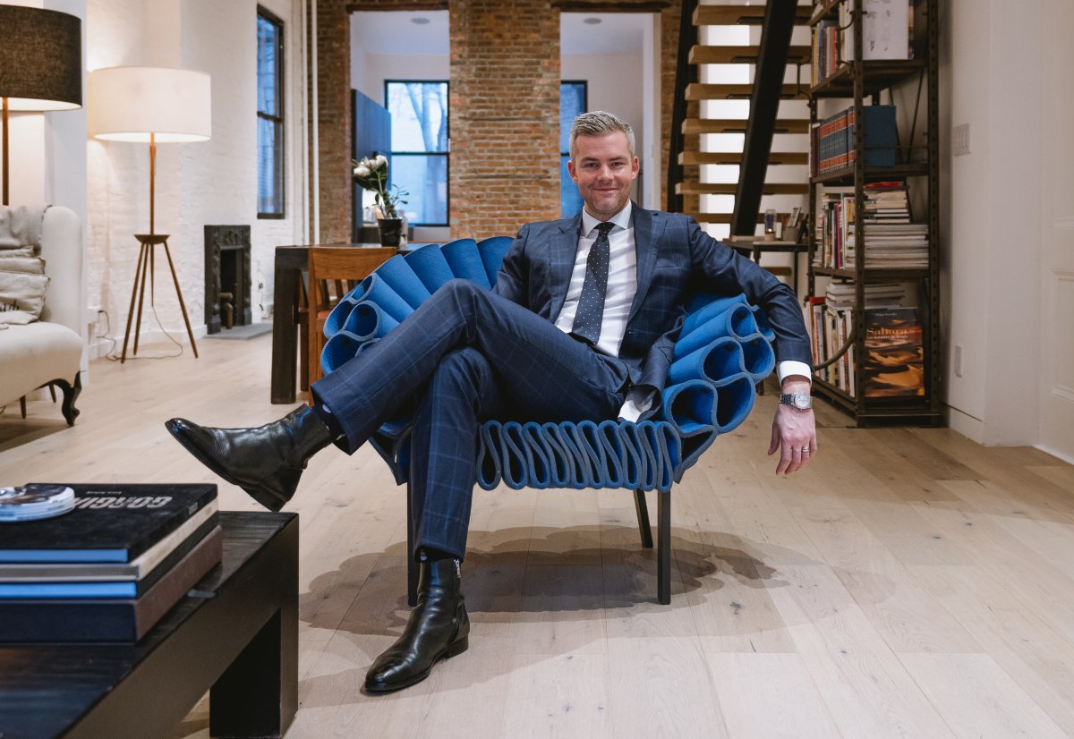 Ryan Serhant looking happy and resting on a vibrant blue lounge chair in a blue suit in a loft space