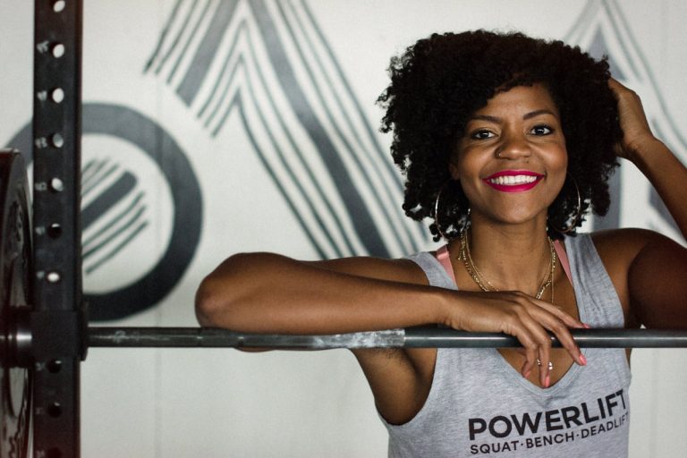 A Black woman smiles at the camera while standing at a squat rack. The woman is Chrissy King.