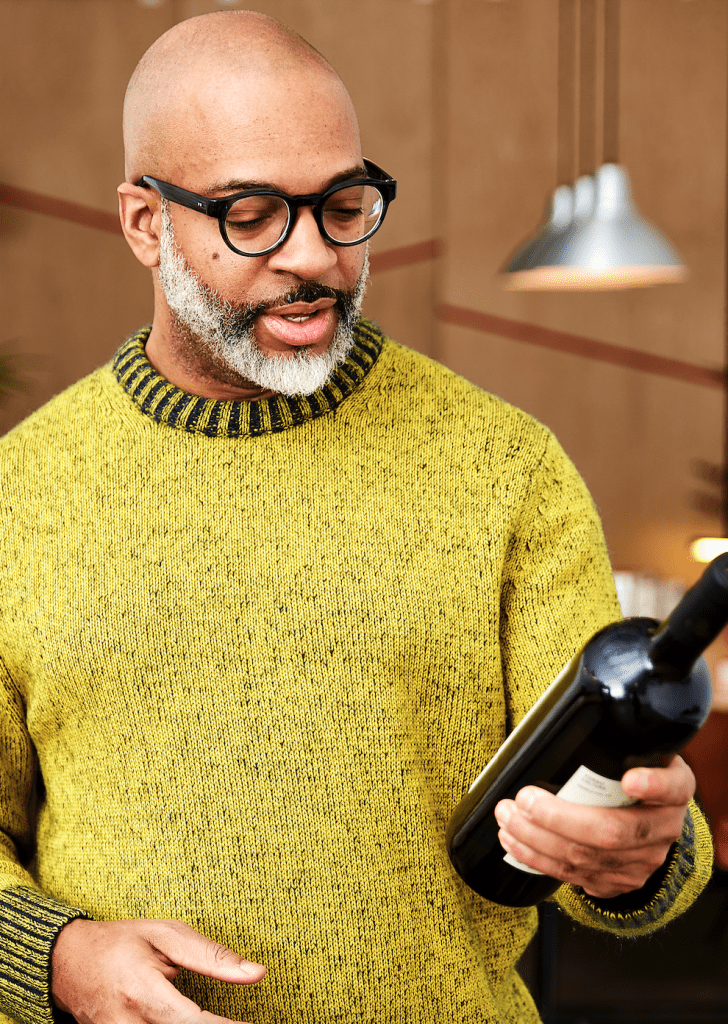 Mean in glasses wearing chartreuse sweater looking down at a bottle of wine.