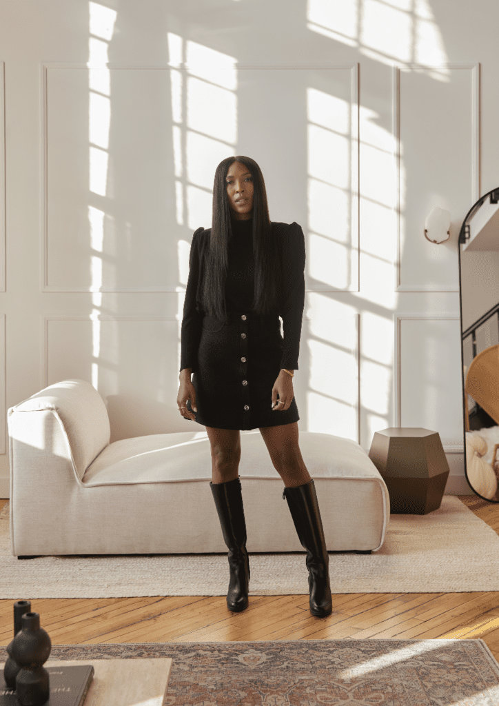 Sasha Exeter with long straight hair standing in a black blouse and skirt with large metal buttons and knee-high black leather boots. She is standing in a living room with a large white loveseat and afternoon light streaming through windows on the wall.