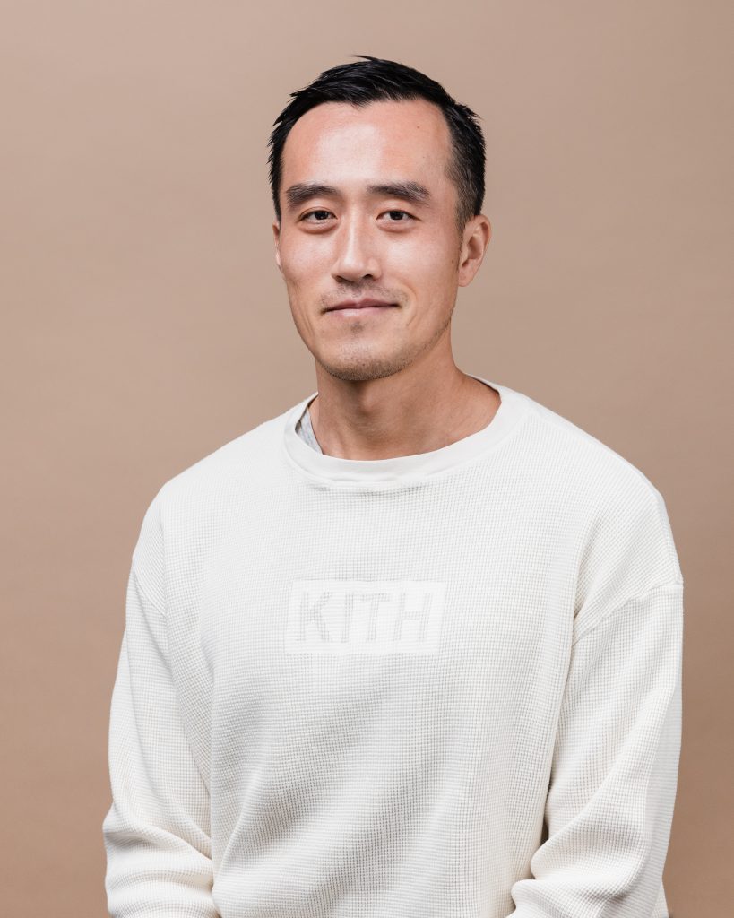 Victor Tam wearing white sweatshirt with words 'KITH' embossed on top against light brown background.