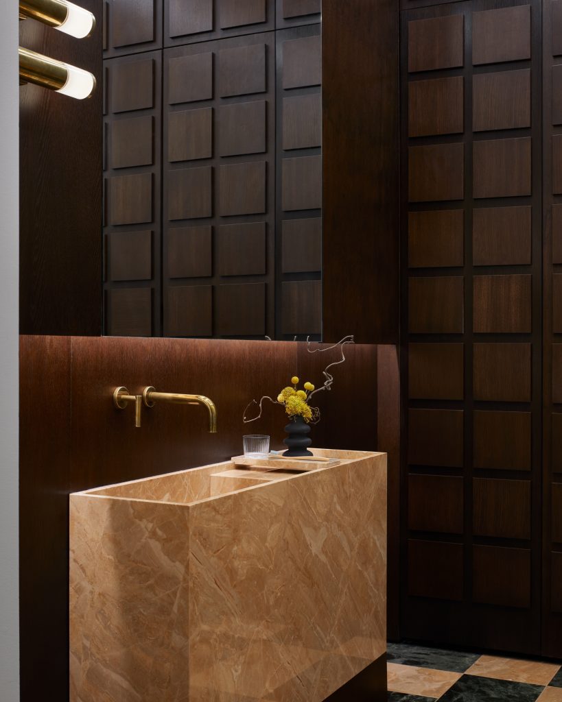 Brown-hued marble sink with brass fixtures featured alongside dark wood square tiling.