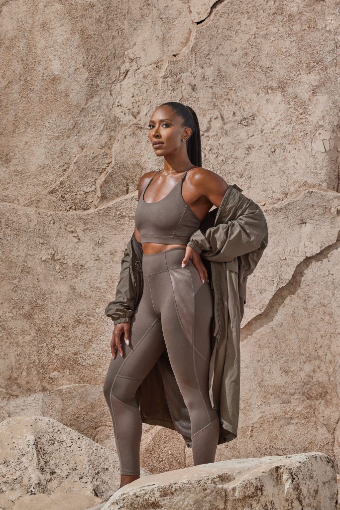 Sasha Exeter posing with hand on hip in a taupe activewear two-piece and jacket against a rocky background
