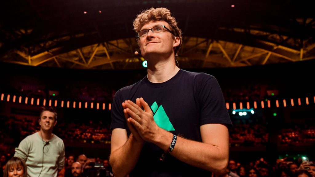 Paddy Cosgrave wearing a black t-shirt with the web summit logo. He is holding his hands in a prayer position and looking up onto a stage with an auditorium of people behind him.