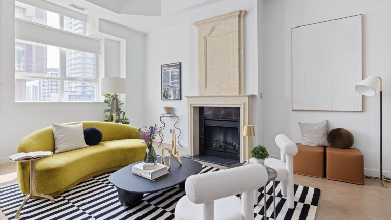 Industrial light-filled loft with a marble fireplace mantle. There is a green sofa situated in front of a black coffee table and white arm chair. They are on top of a black and white rug.
